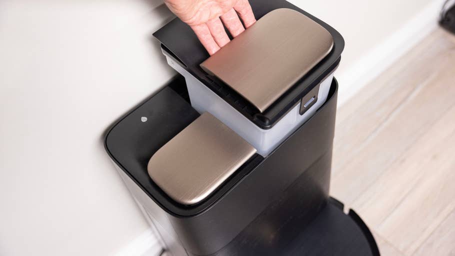 Eine Hand greift nach dem Deckel eines schwarz-silbernen Behälters auf dem Boden.