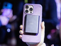 Eine Person hält ein Smartphone mit einem schwarzen Zubehörteil auf der Rückseite in einer überfüllten Umgebung.