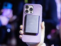 Eine Person hält ein Smartphone mit einem schwarzen Zubehörteil auf der Rückseite in einer überfüllten Umgebung.