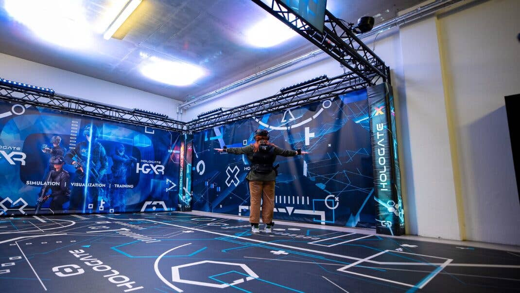 Eine Person mit VR-Ausrüstung steht in einem Hologate-Simulationsraum mit digitalen Grafiken an Wänden und Boden. Eine Person mit VR-Ausrüstung steht in einem Hologate-Simulationsraum mit digitalen Grafiken an Wänden und Boden.