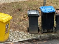 Vier Abfallbehälter in einer Reihe: gelb, schwarz, blau und braun, auf einem Kiesweg mit Gras.