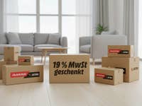 Pakete mit dem Logo von MediaMarkt/Saturn liegen in einem modernen Wohnzimmer auf dem Boden. In der Mitte ist ein Packet mit dem Aufdruck 19 % MwSt geschenkt.