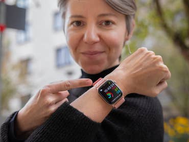 Eine Frau in einem schwarzen Pullover zeigt auf ihre Apple Watch, die Uhrzeit und Aktivitätsdaten anzeigt.