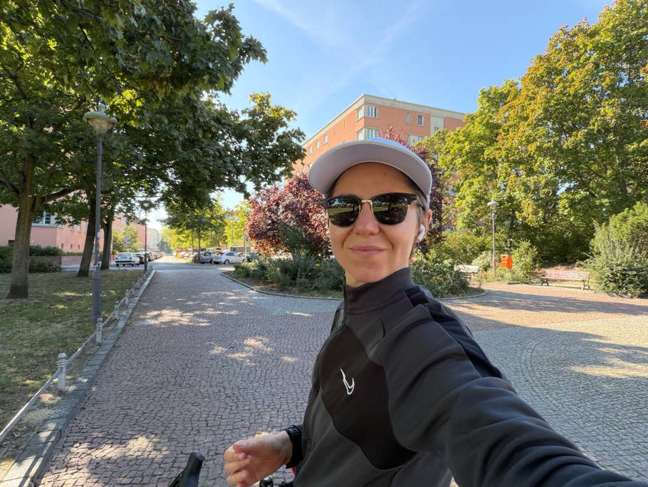 Eine Person in einer schwarzen Jacke und Sonnenbrille lächelt für ein Selfie in einem Park mit Bäumen und Gebäuden im Hintergrund.