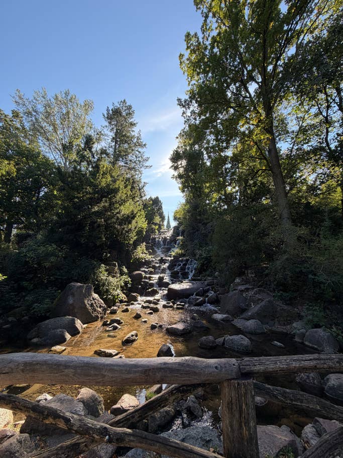 Ein ruhiger Wasserfall fließt durch Steine, umgeben von üppigem Grün unter einem klaren blauen Himmel.