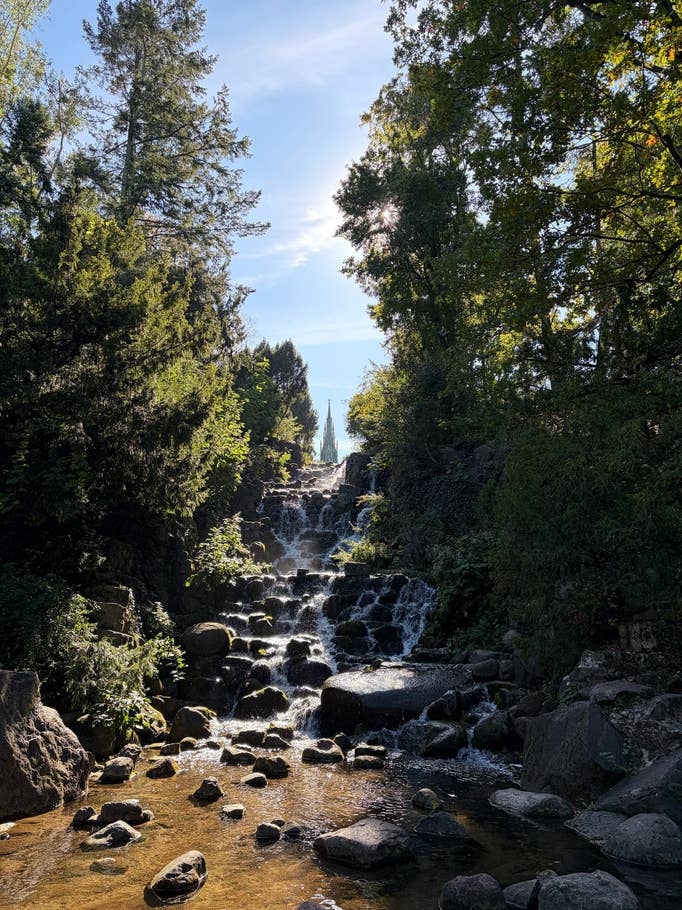 Ein ruhiger Wasserfall fließt über Steine, umgeben von Bäumen unter einem klaren blauen Himmel.