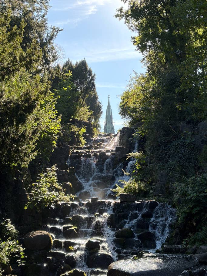 Ein Wasserfall, der über Felsen fließt, umgeben von Grün, mit einem Turm im Hintergrund vor einem blauen Himmel.