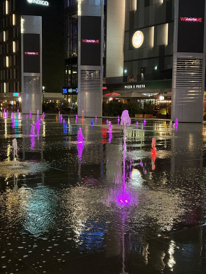 Brunnen mit bunten Lichtern in einem Platz bei Nacht, mit einem 'WILLKOMMEN'-Schild.