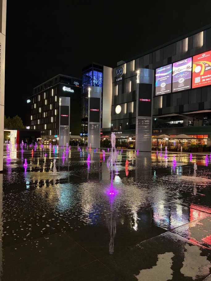 Nachtansicht eines Platzes mit bunten Brunnen und modernen Gebäuden.