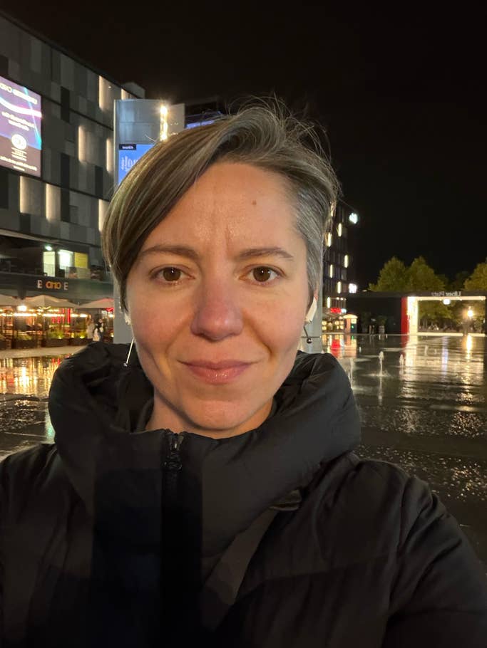 Eine Person mit kurzen Haaren, die eine schwarze Jacke trägt und nachts im Freien in der Nähe beleuchteter Gebäude steht.