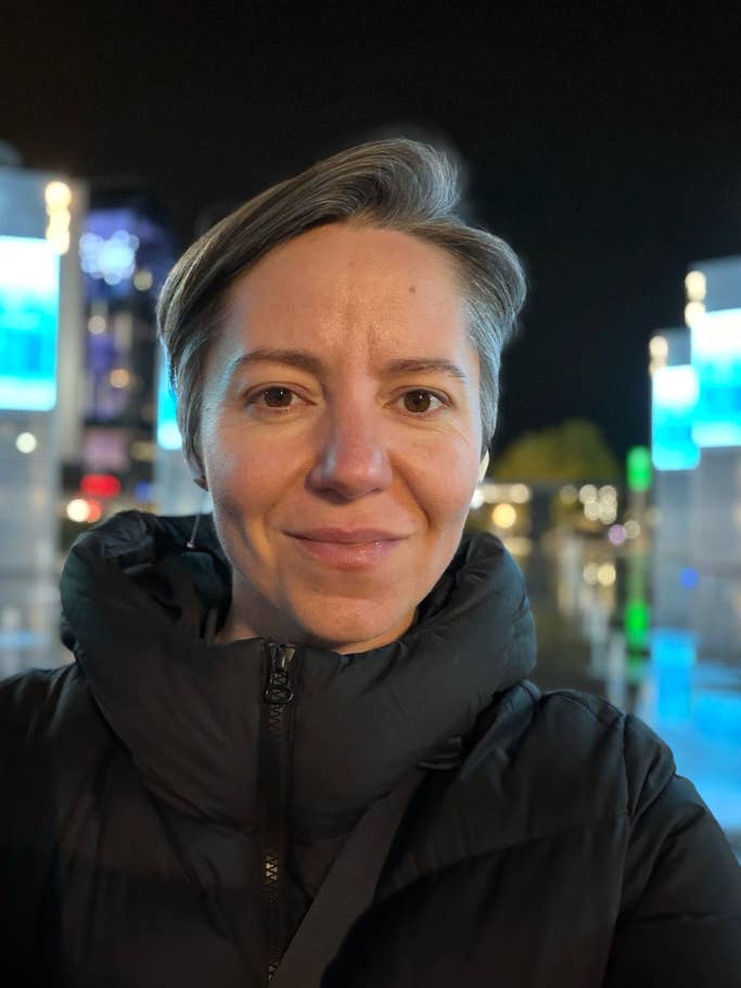 Eine Frau mit kurzem grauem Haar, die eine schwarze Daunenjacke trägt, lächelt nachts mit beleuchteten Gebäuden im Hintergrund.