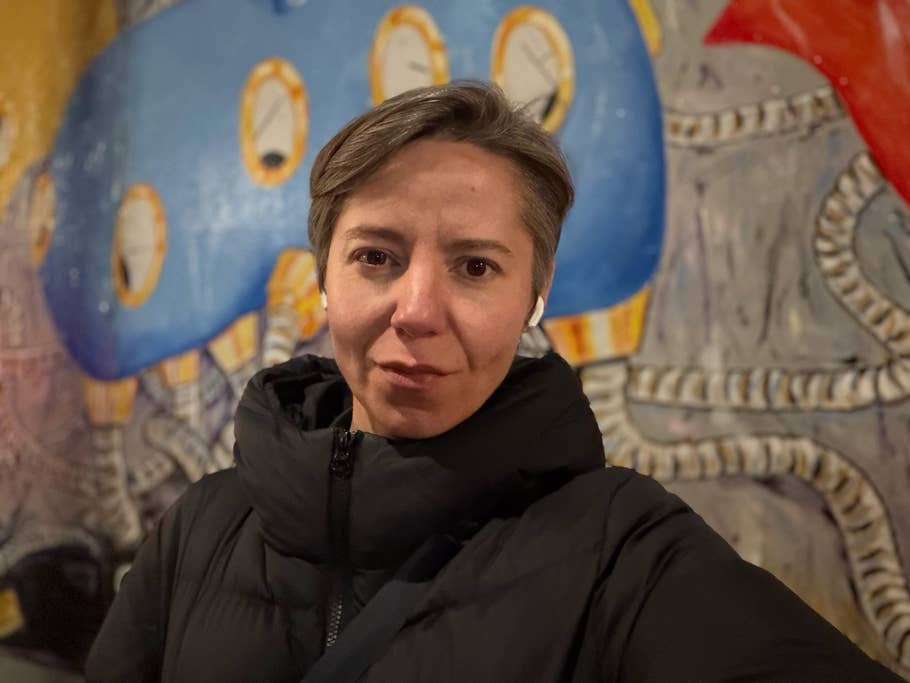 Eine Person mit kurzen Haaren in einer schwarzen Jacke steht vor einem bunten Wandgemälde.