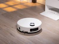 Ein weißer Roboterstaubsauger auf einem Holzboden, mit einer Ladestation im Hintergrund.