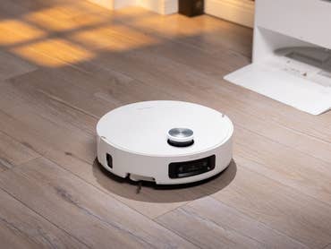 Ein weißer Roboterstaubsauger auf einem Holzboden, mit einer Ladestation im Hintergrund.