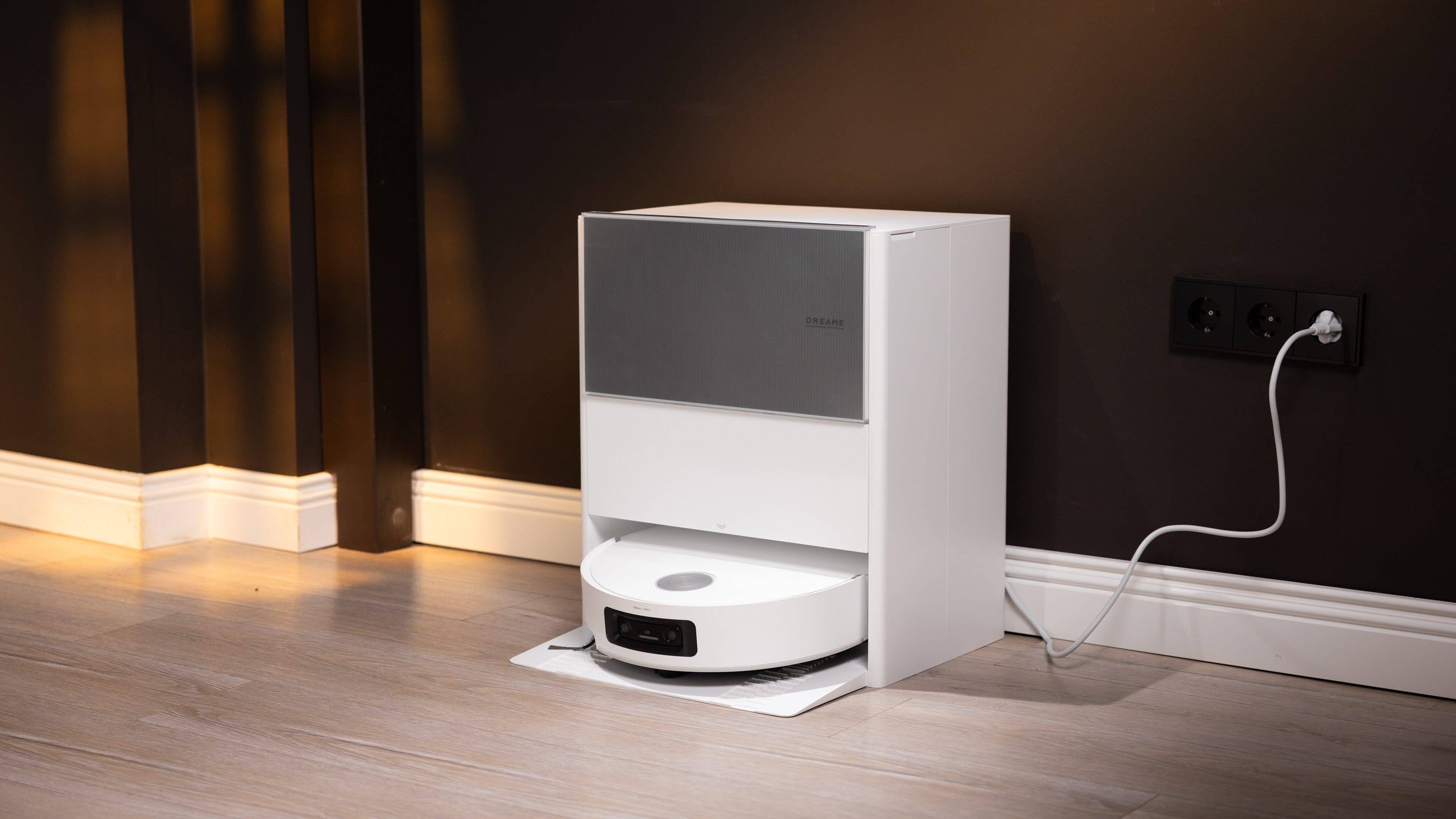 Ein weißer automatischer Staubsauger, angedockt an einer dunklen Wand, mit einem angeschlossenen Stromkabel.