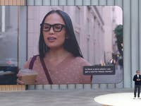 Eine Frau mit Brille fragt nach einem Geschäft in der Nähe, während sie ein Getränk hält. Text: Gibt es ein Geschäft, wo ich eine Karte für Kelsey in der Nähe bekommen kann?