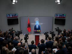 Eine Pressekonferenz in Albanien mit einem Sprecher am Pult, umgeben von Kameras und einer albanischen Flagge.