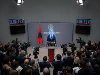 Eine Pressekonferenz in Albanien mit einem Sprecher am Pult, umgeben von Kameras und einer albanischen Flagge.