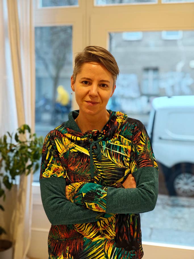 Eine Person mit kurzen Haaren und einem bunten Kapuzenpullover steht mit verschränkten Armen da.