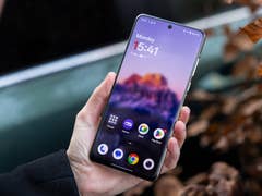 Eine Person hält ein OnePlus 13-Smartphone, das den Startbildschirm mit Uhrzeit und Apps anzeigt.