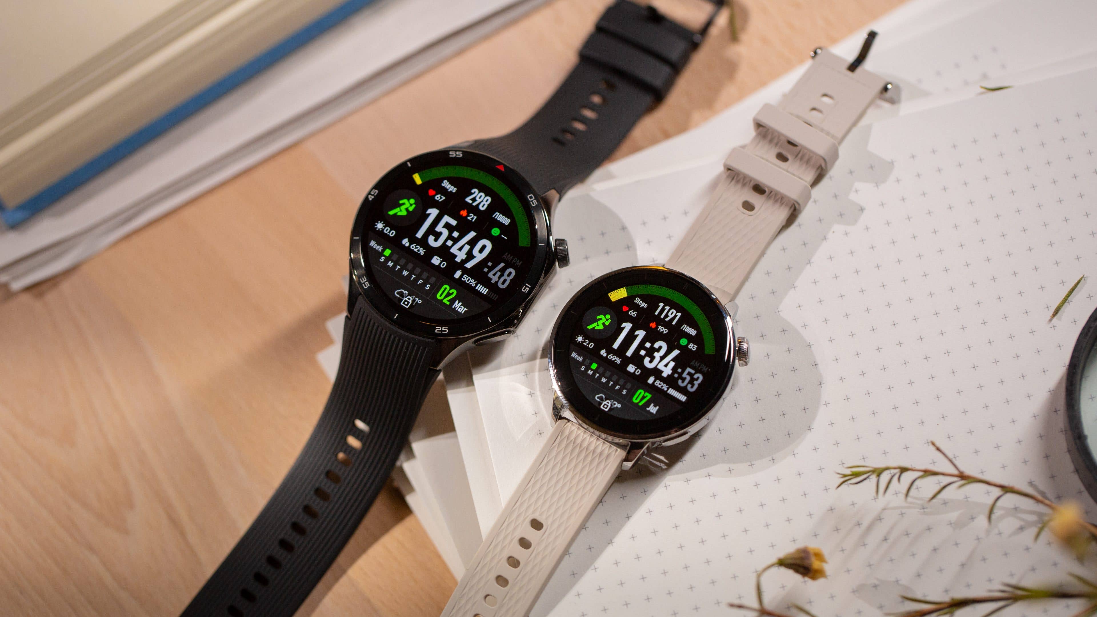 Two OnePlus Watch 3 smartwatches on a desk, one black and one light beige, displaying fitness tracking data.