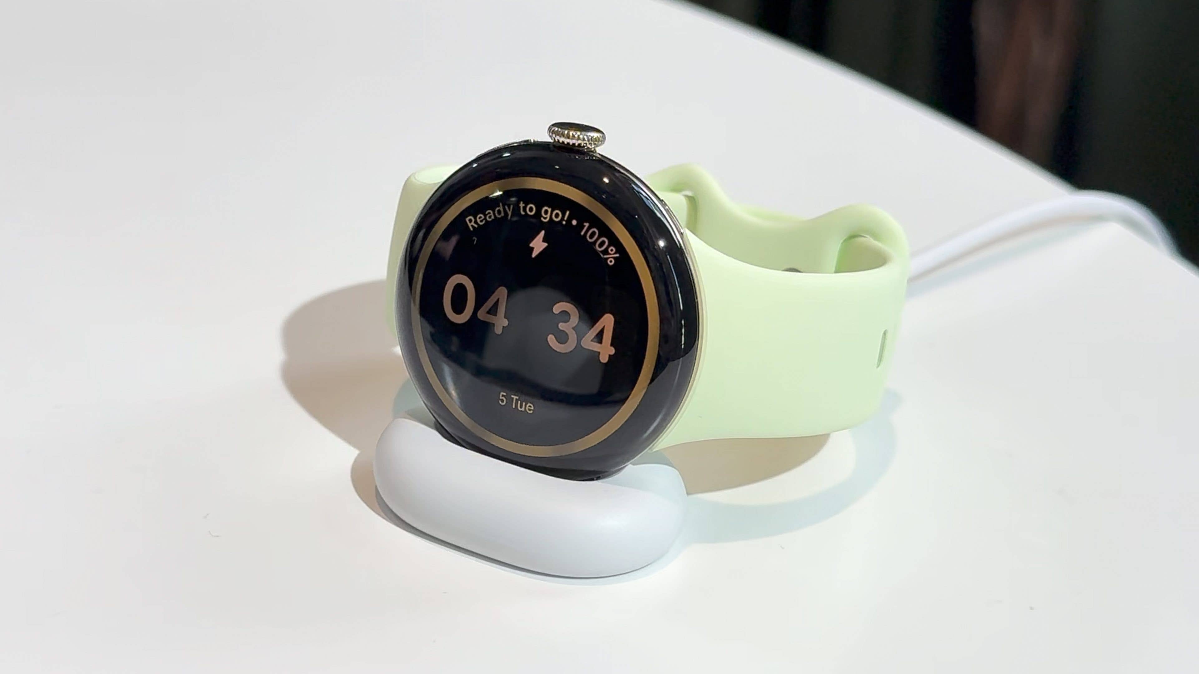 Eine Google Pixel Watch 4 auf einer Ladestation, die die Uhrzeit 04:34 und einen Akkustatus von 100% anzeigt.