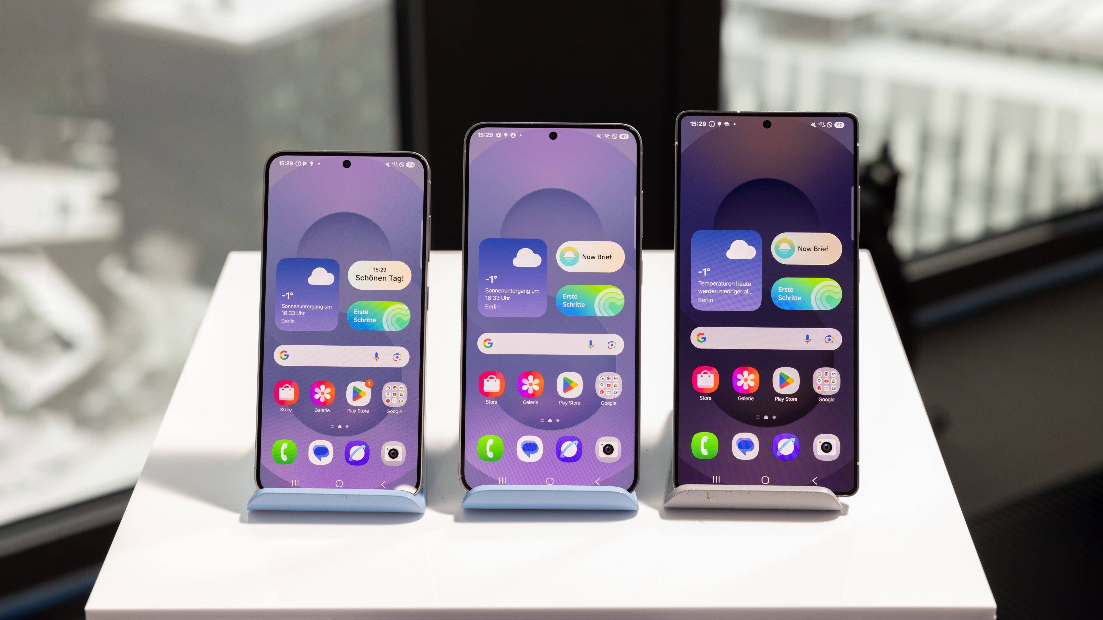 Drei Samsung Galaxy S25-Handys auf Ständern, die verschiedene Startbildschirm-Layouts zeigen.
