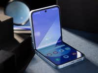 Ein offenes Samsung Galaxy Z Flip 6 Smartphone zeigt seinen Bildschirm mit verschiedenen App-Symbolen und einem blauen Hintergrund.