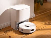 Ein weißer Staubsauger-Roboter neben einer Ladestation auf Holzboden.