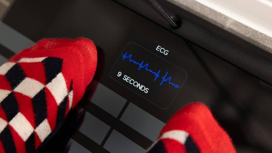 Füße in bunten Socken auf einer Digitalwaage mit den Anzeigen „EKG“ und „9 SEKUNDEN“.