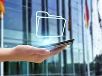 Eine Hand hält ein Smartphone mit einem schwebenden Ordnersymbol vor einem modernen Gebäude.