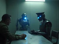 Ein humanoider Roboter verhört einen Mann in einem schwach beleuchteten Raum, während ein anderer Roboter daneben steht.