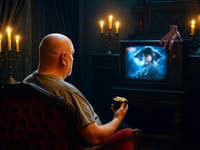 Ein glatzköpfiger Mann sitzt in einem Sessel und schaut sich eine gruselige Szene im Fernsehen an, während Kerzen den dunklen Raum beleuchten.