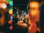 Eine Hand hält ein Smartphone, das eine hell erleuchtete Gasse zeigt.