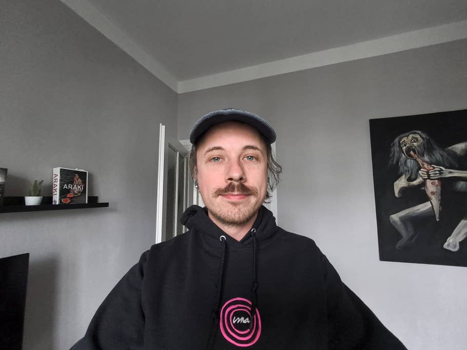 Eine Person mit Schnurrbart trägt einen schwarzen Hoodie und steht in einem Raum mit einem Gemälde und Büchern.