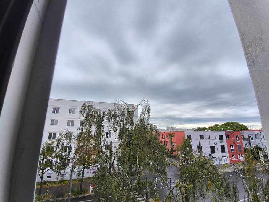 Blick auf einen bewölkten Himmel und Stadtgebäude durch ein Fenster mit Bäumen im Vordergrund.