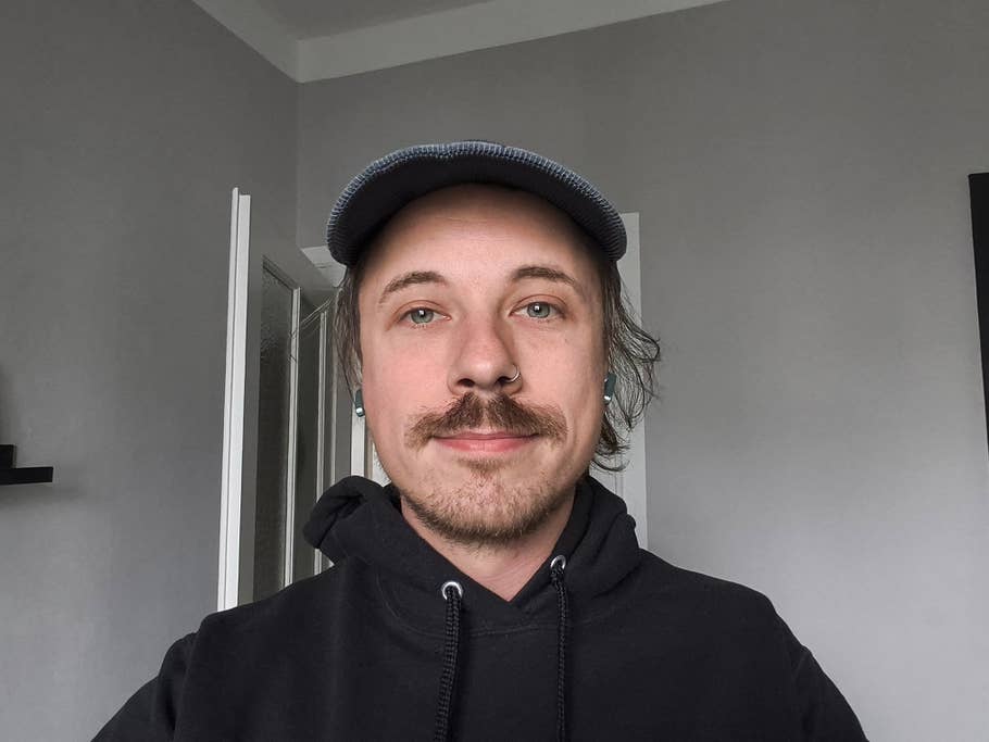 Eine Person mit Schnurrbart und Mütze, die einen schwarzen Hoodie trägt, steht in einem Raum mit grauen Wänden.