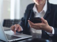 Eine Frau sitzt am Laptop und schaut auf ihr Handy