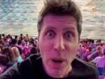 Sam Altman von OpenAI macht ein Selfie in einer überfüllten Arena während eines Schwimmwettbewerbs, mit bunten Lichtern im Publikum.