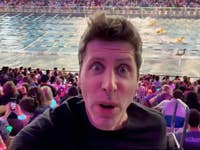 Sam Altman von OpenAI macht ein Selfie in einer überfüllten Arena während eines Schwimmwettbewerbs, mit bunten Lichtern im Publikum.