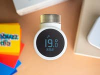 Ein weißes Smart-Thermostat zeigt 19,0 °C und 26,8 °C auf einer beigen Oberfläche mit bunten Karten im Hintergrund an.