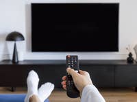 Eine Person hält eine Fernbedienung vor einem schwarzen Smart-TV.