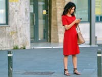Eine Frau im roten Kleid steht auf dem Bürgersteig und schaut auf ihr Handy