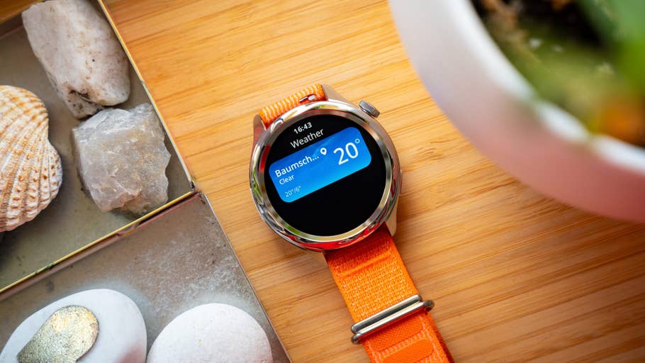 Eine Xiaomi Watch S4 zeigt Wetterinformationen mit einem orangefarbenen Armband, umgeben von Steinen und einer Pflanze.