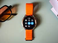 Eine Xiaomi Watch S4 mit orangefarbenem Armband, die verschiedene App-Symbole anzeigt, neben Sonnenbrillen und Zeitschriften.