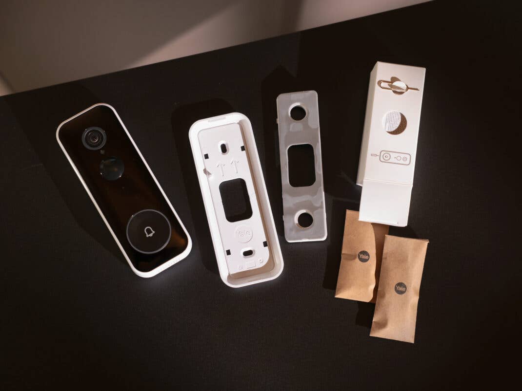 Die demontierte Yale Video-Türklingel liegt mit Halterungen, Montagematerial und kleinen braunen Verpackungstüten auf einer dunklen Oberfläche.